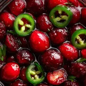 A spoonful of sweet and spicy candied jalapeños with cranberries being lifted from a glass jar. The syrup drips off the spoon, highlighting the delicious texture.