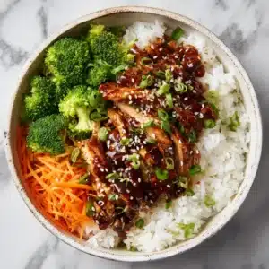 An overhead view of a homemade teriyaki chicken bowl with broccoli, carrots, and rice, garnished with sesame seeds.