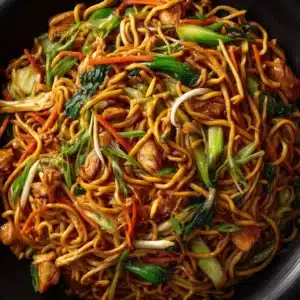 A close-up shot of the chow mein ingredients being stir-fried in a hot wok, with steam rising from the fresh vegetables and noodles.