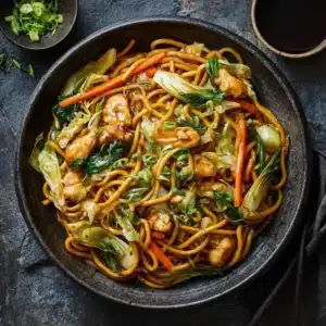 A close-up view of chow mein noodles being stir-fried in a wok with chicken and crisp vegetables like carrots and cabbage.