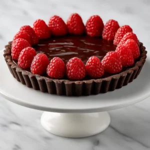 A whole chocolate raspberry tart shown from a three-quarters angle, resting on a marble surface. The rich chocolate ganache filling is perfectly set inside the dark chocolate crust.