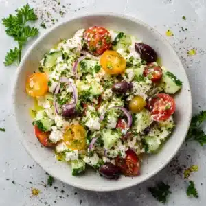 A close-up shot of the fresh ingredients for a Greek cottage cheese salad, including cherry tomatoes, diced cucumber, and red onion in a bowl.