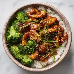 An overhead view of a homemade teriyaki chicken bowl with fresh broccoli, carrots, and fluffy white rice in a ceramic bowl.