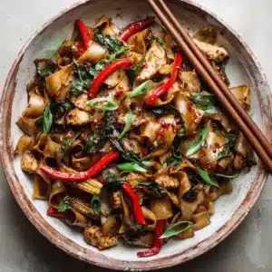 Fresh ingredients for Thai Drunken Noodles including red bell pepper, baby corn, Thai basil, scallions, wide rice noodles, and chicken breast pieces.