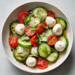 A minimalist white bowl sitting on a plain light grey surface containing fresh Cucumber Mozzarella Salad with coarse sea salt flakes and black pepper.