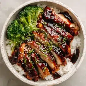 A close-up, overhead shot of a Teriyaki Chicken Rice Bowl. Slices of pan-seared chicken are coated in a thick, glossy, dark brown sauce and garnished with sesame seeds and scallions.