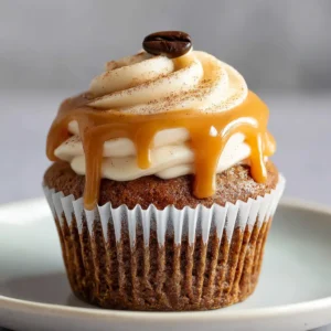 A close-up of a single Gingerbread Caramel Latte Cupcake in a white liner. It features a dark gingerbread sponge, tall swirl of latte buttercream, caramel drizzle, and a coffee bean garnish.