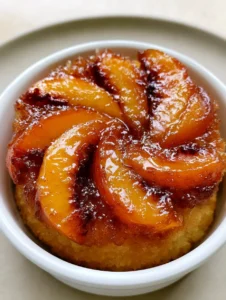 Individual mini peach upside-down cake in a white ramekin, featuring golden-orange peach slices and dark amber edges. (Mini Peach Upside-Down Cake)