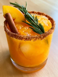Close up of a pulpy Fall Peach Cocktail with large clear ice cubes and a thick yellow peach wedge garnish.