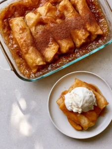 A clear glass rectangular baking dish filled with golden-brown flour tortillas rolled with peaches, drenched in bubbling amber-colored cinnamon butter syrup.